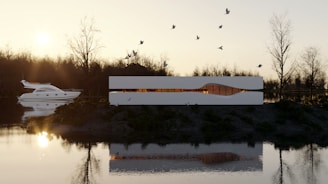 A modern white building with a sleek design sits by a calm river, surrounded by barren trees. The sunset casts warm light, contrasting with the building's minimalistic architecture. Birds are flying above, and a luxury yacht is seen on the water.