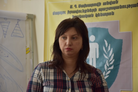 A person with dark hair wearing a plaid shirt stands in front of a poster with text in another language. There is a flip chart on the left with sketches or drawings.