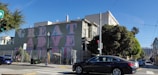 A mural on the side of a building has the words 'GREAT ADVENTURE' in large, colorful letters. The street is lined with trees, and a black car is driving past. The scene includes a streetlight, a traffic light with a red signal, and several buildings, including a Victorian-style structure. The sky is clear and blue.