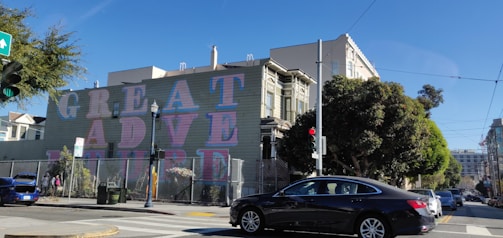 A mural on the side of a building has the words 'GREAT ADVENTURE' in large, colorful letters. The street is lined with trees, and a black car is driving past. The scene includes a streetlight, a traffic light with a red signal, and several buildings, including a Victorian-style structure. The sky is clear and blue.