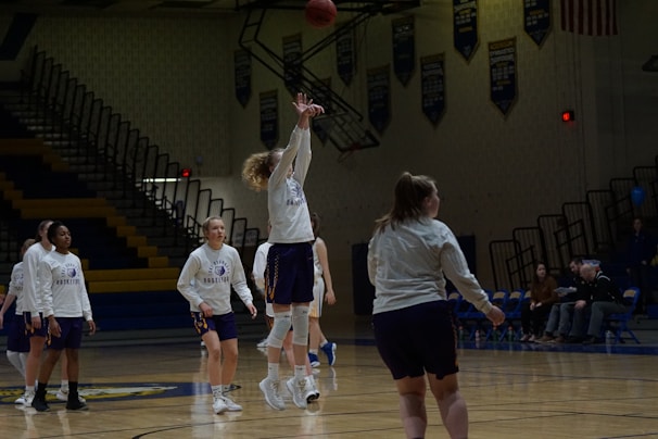 Several basketball players in matching uniforms practice on a dimly lit gymnasium court. One player is jumping to shoot the ball towards a hoop that is not visible in the frame. Other players are either standing or walking around the court. Spectators are seated on the bleachers in the background.