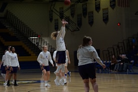 Several basketball players in matching uniforms practice on a dimly lit gymnasium court. One player is jumping to shoot the ball towards a hoop that is not visible in the frame. Other players are either standing or walking around the court. Spectators are seated on the bleachers in the background.