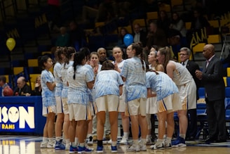 A group of female basketball players huddled together on a basketball court, wearing white uniforms with blue accents. Coaches or team staff stand nearby, and a crowd is visible in the background seated in bleachers.