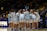 A group of female basketball players huddled together on a basketball court, wearing white uniforms with blue accents. Coaches or team staff stand nearby, and a crowd is visible in the background seated in bleachers.
