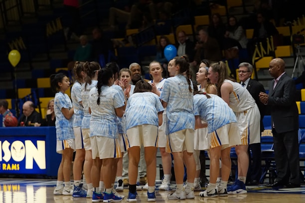 A group of female basketball players huddled together on a basketball court, wearing white uniforms with blue accents. Coaches or team staff stand nearby, and a crowd is visible in the background seated in bleachers.