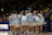 A group of female basketball players huddled together on a basketball court, wearing white uniforms with blue accents. Coaches or team staff stand nearby, and a crowd is visible in the background seated in bleachers.