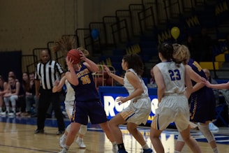 A women's basketball game where one player in a dark jersey is holding the ball tightly, while another player in a white jersey is attempting to block her. A referee stands nearby, observing the game. The backdrop consists of empty bleachers with some banners and balloons in the background.