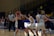 A women's basketball game where one player in a dark jersey is holding the ball tightly, while another player in a white jersey is attempting to block her. A referee stands nearby, observing the game. The backdrop consists of empty bleachers with some banners and balloons in the background.