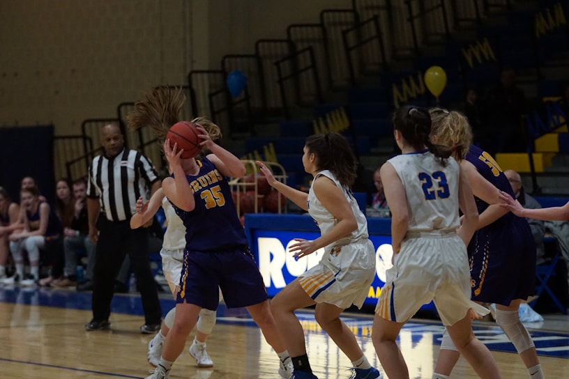 A women's basketball game where one player in a dark jersey is holding the ball tightly, while another player in a white jersey is attempting to block her. A referee stands nearby, observing the game. The backdrop consists of empty bleachers with some banners and balloons in the background.