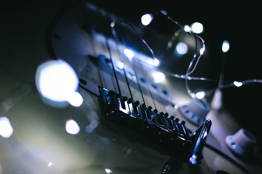 A close-up view of an electric guitar decorated with string lights, creating a moody ambiance. The focus is on the guitar's strings and bridge, with the lights casting a soft glow.