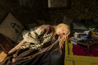 A person with long blonde hair is asleep, resting their head on a bright yellow table. They are draped in a patchwork blanket featuring various patterns, including leopard print. Surrounding them are several decorative pillows with artistic designs, creating a cozy setting. Various items such as a bowl, containers, and art supplies are scattered on the table, giving a sense of casual disarray. The setting is dimly lit with warm sunlight streaming in, casting soft shadows.
