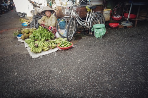 A street vendor is sitting on the ground beside a bicycle, surrounded by a variety of fruits and vegetables, including bananas, limes, and leafy greens. The vendor wears a conical hat and is in an outdoor market setting. The scene includes baskets and other market items laid out on the pavement.