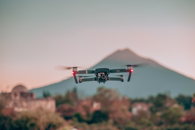 A drone is hovering in the air with blurred landscape in the background. The backdrop includes a mountain with a soft gradient of colors in the sky, transitioning from a pinkish hue to a light blue. The foreground features a mix of greenery and some out-of-focus buildings.