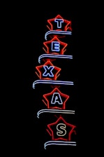 Neon light sign spelling 'TEXAS', with each letter enclosed in a star-shaped border. The stars are outlined in bright red, and the letters are illuminated in blue. The background is completely black, highlighting the vibrant neon colors.