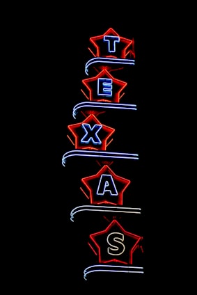 Neon light sign spelling 'TEXAS', with each letter enclosed in a star-shaped border. The stars are outlined in bright red, and the letters are illuminated in blue. The background is completely black, highlighting the vibrant neon colors.