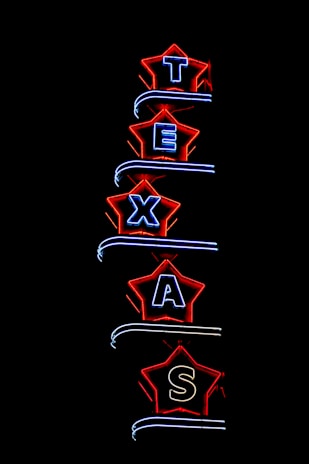 Neon light sign spelling 'TEXAS', with each letter enclosed in a star-shaped border. The stars are outlined in bright red, and the letters are illuminated in blue. The background is completely black, highlighting the vibrant neon colors.