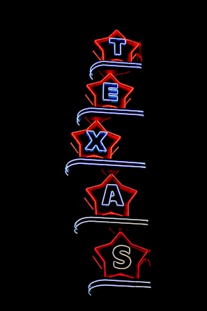Neon light sign spelling 'TEXAS', with each letter enclosed in a star-shaped border. The stars are outlined in bright red, and the letters are illuminated in blue. The background is completely black, highlighting the vibrant neon colors.