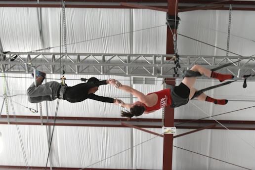 Two individuals are performing a mid-air trapeze maneuver inside a gymnasium. One person is suspended upside down, wearing a red tank top and grey shorts, reaching out to the other, who is dressed in a black shirt and grey pants with harnesses securing them to the overhead structure.