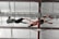 Two individuals are performing a mid-air trapeze maneuver inside a gymnasium. One person is suspended upside down, wearing a red tank top and grey shorts, reaching out to the other, who is dressed in a black shirt and grey pants with harnesses securing them to the overhead structure.