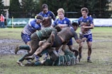 A rugby match taking place on a muddy field with players wearing muddy uniforms. A group of athletes in blue and green jerseys are engaged in a scrummage as they compete for the ball.