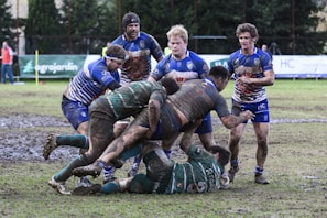 A rugby match taking place on a muddy field with players wearing muddy uniforms. A group of athletes in blue and green jerseys are engaged in a scrummage as they compete for the ball.