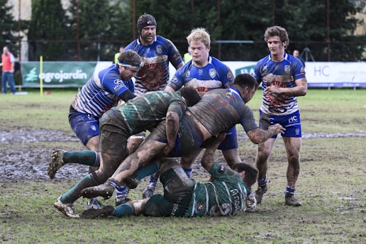 A rugby match taking place on a muddy field with players wearing muddy uniforms. A group of athletes in blue and green jerseys are engaged in a scrummage as they compete for the ball.