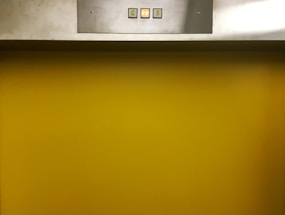 A metal panel with a display showing buttons labeled G, 2, and 3 is mounted above a bright yellow background. The buttons are small and square, containing numbers or letters that indicate different levels.