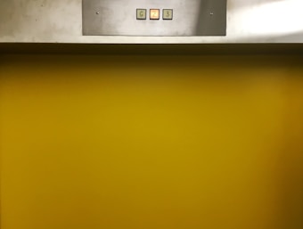 A metal panel with a display showing buttons labeled G, 2, and 3 is mounted above a bright yellow background. The buttons are small and square, containing numbers or letters that indicate different levels.
