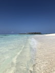 Clear turquoise waves gently lap against a pristine sandy beach under a vast, cloudless blue sky. In the distance, a line of overwater bungalows extends over the ocean, creating a serene and picturesque tropical setting.