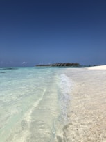 Clear turquoise waves gently lap against a pristine sandy beach under a vast, cloudless blue sky. In the distance, a line of overwater bungalows extends over the ocean, creating a serene and picturesque tropical setting.
