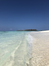 Clear turquoise waves gently lap against a pristine sandy beach under a vast, cloudless blue sky. In the distance, a line of overwater bungalows extends over the ocean, creating a serene and picturesque tropical setting.
