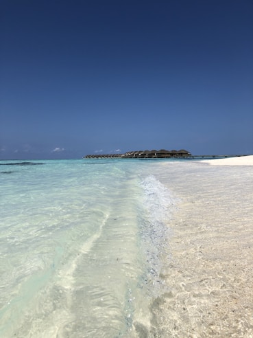 Clear turquoise waves gently lap against a pristine sandy beach under a vast, cloudless blue sky. In the distance, a line of overwater bungalows extends over the ocean, creating a serene and picturesque tropical setting.