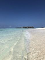 Clear turquoise waves gently lap against a pristine sandy beach under a vast, cloudless blue sky. In the distance, a line of overwater bungalows extends over the ocean, creating a serene and picturesque tropical setting.