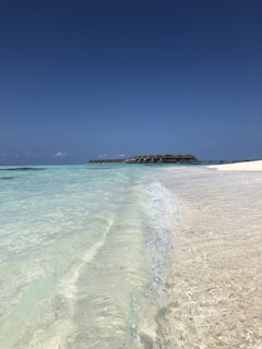 Clear turquoise waves gently lap against a pristine sandy beach under a vast, cloudless blue sky. In the distance, a line of overwater bungalows extends over the ocean, creating a serene and picturesque tropical setting.