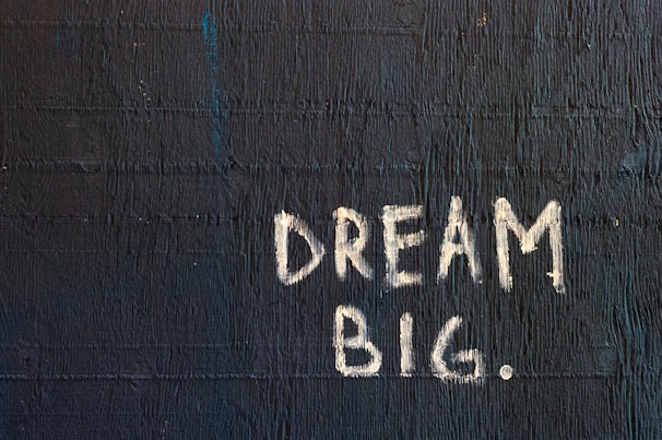 The image features the phrase 'DREAM BIG.' written in white chalk-like lettering on a textured, dark blue wall. The text stands out sharply against the rough surface, which exhibits horizontal linear patterns.