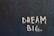 The image features the phrase 'DREAM BIG.' written in white chalk-like lettering on a textured, dark blue wall. The text stands out sharply against the rough surface, which exhibits horizontal linear patterns.