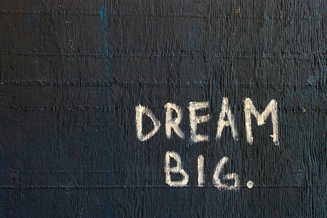 The image features the phrase 'DREAM BIG.' written in white chalk-like lettering on a textured, dark blue wall. The text stands out sharply against the rough surface, which exhibits horizontal linear patterns.