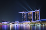 A nighttime cityscape featuring a large, brightly lit hotel with three towers connected by an illuminated platform at its top. Spotlights beam into the sky, and their reflections shimmer on the calm water below. To the left, a modern, lotus-shaped building adds a touch of unique architecture to the scene.