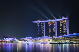 A nighttime cityscape featuring a large, brightly lit hotel with three towers connected by an illuminated platform at its top. Spotlights beam into the sky, and their reflections shimmer on the calm water below. To the left, a modern, lotus-shaped building adds a touch of unique architecture to the scene.
