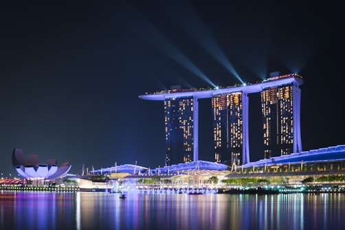 A nighttime cityscape featuring a large, brightly lit hotel with three towers connected by an illuminated platform at its top. Spotlights beam into the sky, and their reflections shimmer on the calm water below. To the left, a modern, lotus-shaped building adds a touch of unique architecture to the scene.