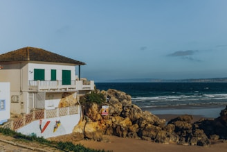A coastal scene featuring a white house with green shutters situated on rocky terrain by the ocean. The house is positioned near the edge of a sandy beach with gentle waves lapping at the shore. Vibrant artwork decorates the building's wall, and the area is surrounded by rocky outcrops and sparse vegetation. The sky is clear with a few scattered clouds.
