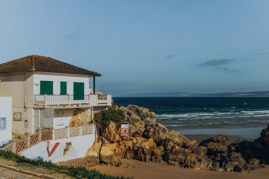 A coastal scene featuring a white house with green shutters situated on rocky terrain by the ocean. The house is positioned near the edge of a sandy beach with gentle waves lapping at the shore. Vibrant artwork decorates the building's wall, and the area is surrounded by rocky outcrops and sparse vegetation. The sky is clear with a few scattered clouds.
