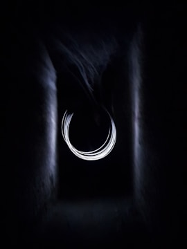 A dark tunnel-like structure with a central circular light pattern that appears abstract and ethereal. The light swirls in a concentric manner, creating an illusion of a portal or vortex.