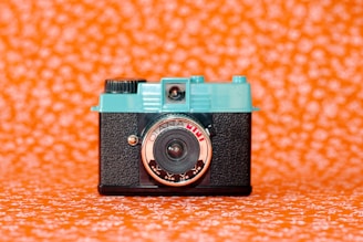 A vintage-style camera with a turquoise top and black textured body is centered against a vivid orange background patterned with white floral designs. The lens is prominently labeled and has a reflective surface.