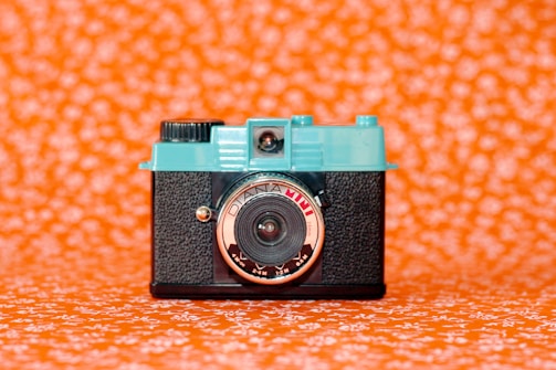 A vintage-style camera with a turquoise top and black textured body is centered against a vivid orange background patterned with white floral designs. The lens is prominently labeled and has a reflective surface.