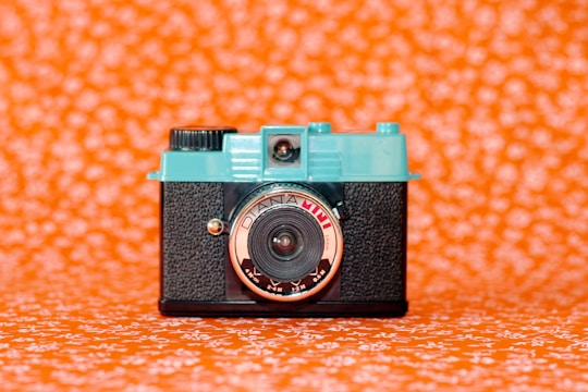A vintage-style camera with a turquoise top and black textured body is centered against a vivid orange background patterned with white floral designs. The lens is prominently labeled and has a reflective surface.
