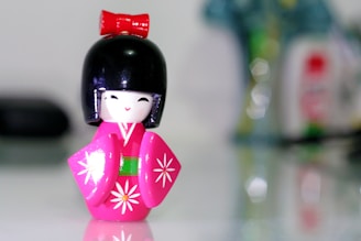 A small, colorful figurine resembling a traditional Japanese doll, known as a Kokeshi, with a black bob hairstyle and a vibrant pink kimono decorated with white floral patterns. The figure has a red bow on top, a cheerful facial expression, and stands against a blurred, indistinct background.