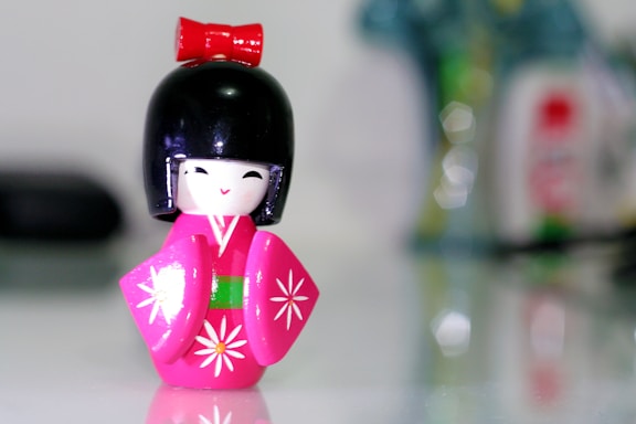 A small, colorful figurine resembling a traditional Japanese doll, known as a Kokeshi, with a black bob hairstyle and a vibrant pink kimono decorated with white floral patterns. The figure has a red bow on top, a cheerful facial expression, and stands against a blurred, indistinct background.