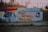 A mural painted on a concrete wall features the words 'Open Streets Detroit' in bold black letters. The background includes colorful abstract designs with shades of blue, red, and yellow. Social media handles are also painted above the text. The cobblestone street and metallic fence in the background add an urban feel.