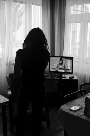 A person stands in front of a television in a dimly lit room. Light filters through curtains from large windows behind the individual. The television displays an image of an athlete with Olympic symbols in the background. The room contains a few pieces of furniture, including a table with a box and a remote on it.